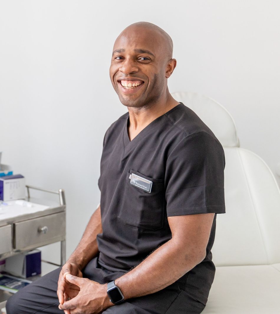 Dr Richard seated in a clinic, smiling in black scrubs